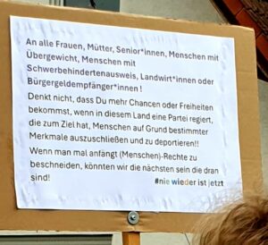 Auf Papier auf einer Papptafel geschrieben steht: An alle Fraune, Mütter, Senior*innen, Menschen mit Übergewicht, Menschen mit Schwerbehindertenausweis, Landwirt*innen oder Bürgergeldempfänger*innen! Denkt nicht, dass Du mehr Chancen oder Freiheiten bekommst, wenn in diesem Land eine Partei regiert, die zum Ziel hat Menschen auf Grund bestimmter Merkmale auszuschließen und zu deportieren!! Wenn man mal anfängt (Menschen-) Rechte zu beschneiden, könnten wir die nächsten sein, die dran sind! #NieWiederIstJetzt