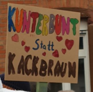 Pappplakat: Kunterbunt statt Kackbraun. Erstes Wort in Bunter Schrift, das letzte in braun. Dazu 7 rote Herzen