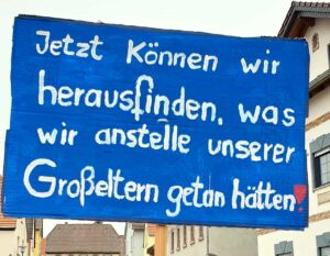 Eine blau bemalte Pappe mit weißer Aufschrift: Jetzt können wir herausfinden, was wir anstelle unserer Großeltern getan hätten.